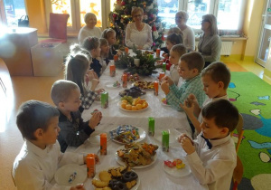 Pani dyrektor Maria Królikowska, pani Agnieszka Piekarska, pani Arleta Kalinowska oraz dzieci jedzą słodki i owocowy poczęstunek.